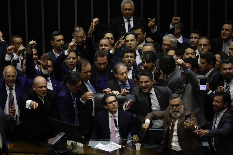 A imagem mostra um grupo de parlamentares em uma sessão do Congresso Nacional. Eles estão em pé, com expressões intensas, alguns levantando os punhos e outros gesticulando. O ambiente é formal, com mesas e equipamentos de votação ao fundo. A maioria dos homens está vestida com ternos e gravatas, e há uma sensação de agitação e debate acalorado entre os presentes.