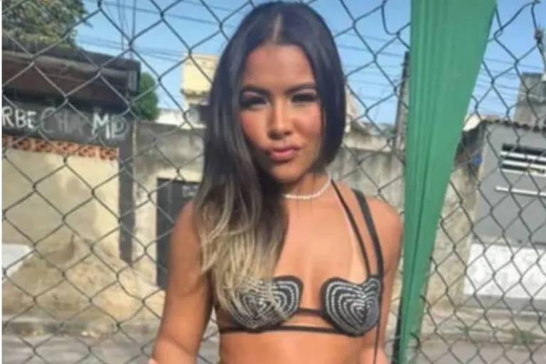 Uma jovem está posando em frente a uma cerca. Ela usa um top preto com detalhes e uma saia de couro preta. A jovem segura um copo e faz um sinal de paz com a mão. Ao fundo, há uma parede e uma estrutura que parecem ser de uma área urbana.