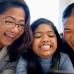 A close up of two women and a girl that look like family, looking at a book or a screen and sharing a laugh.