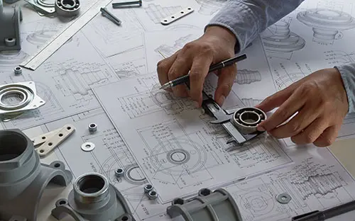 A person measuring mechanical parts with calipers while referencing engineering drawings and specifications